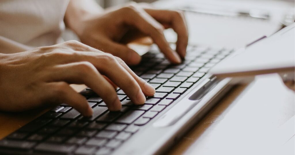 Close-up of hands typing on a black keyboard. The scene is well-lit, showing the fingers actively pressing keys. A part of a laptops open screen is visible, suggesting a work or study setting.
